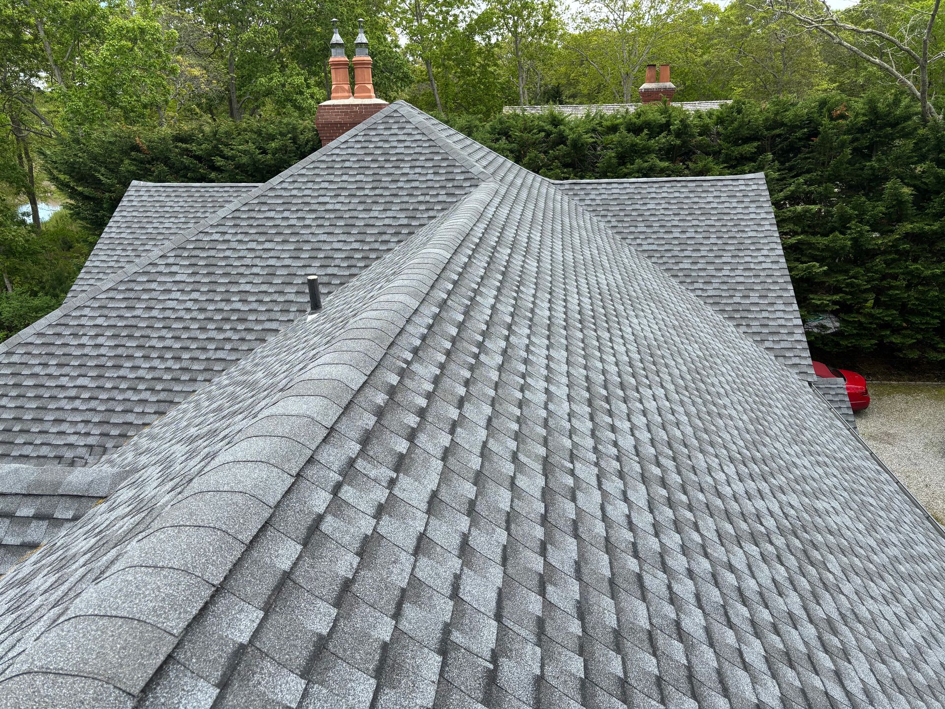 Gray shingled roof of a house set among green trees with two chimneys visible