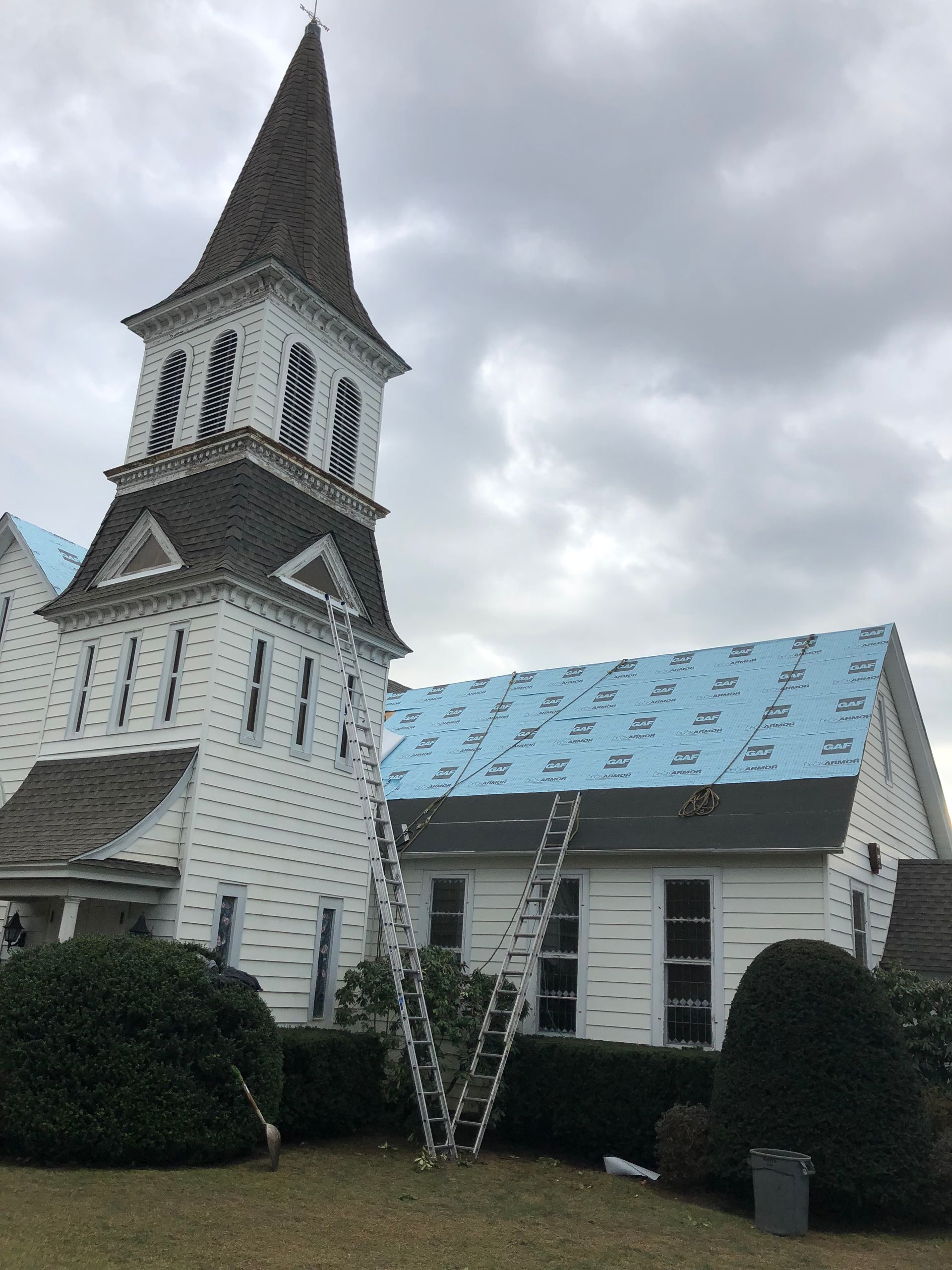 White church building with steeple, ladders against the roof, and blue underlayment