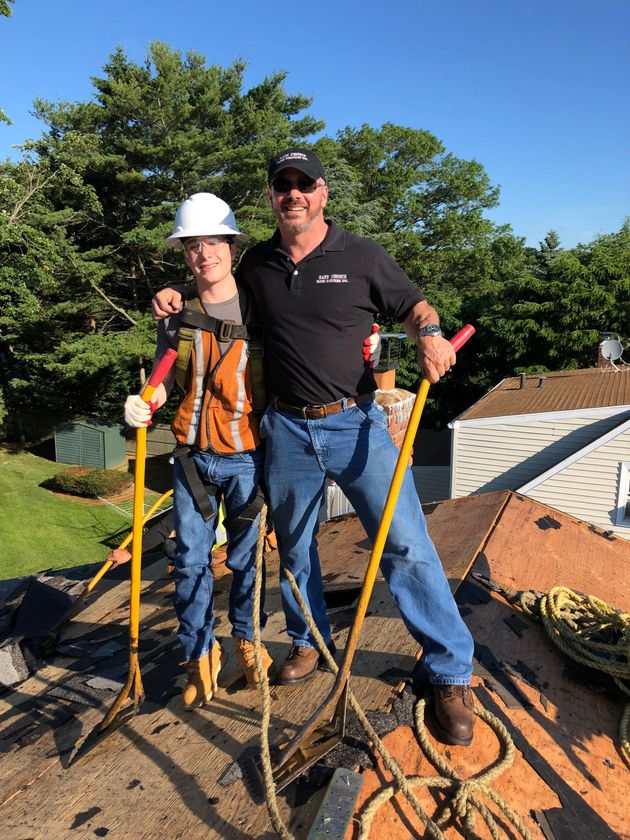 Two people stand on a roof, one in safety gear, holding tools on a sunny day