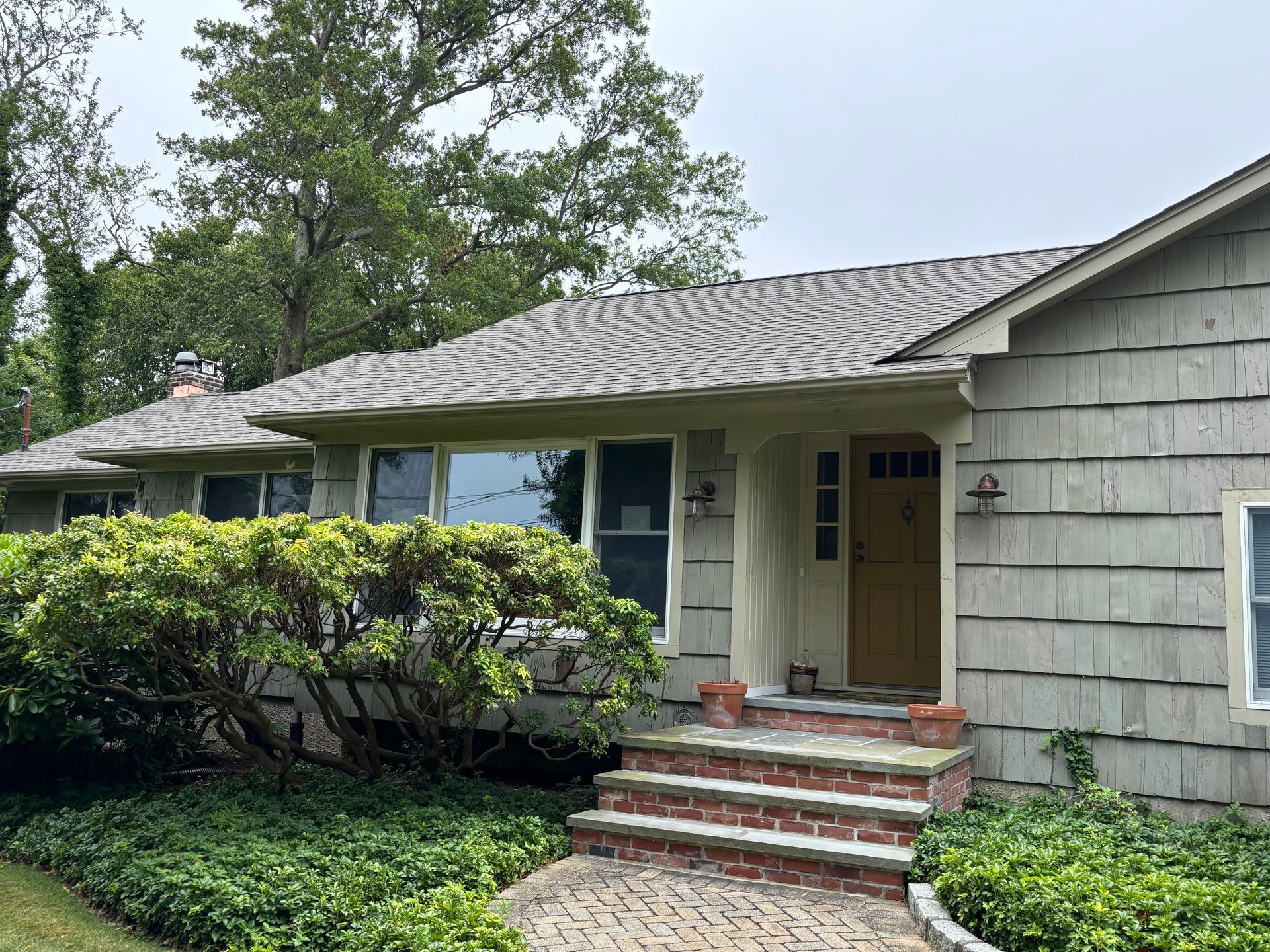 Light gray house with a yellow door, red brick steps, and a lush green bush in front