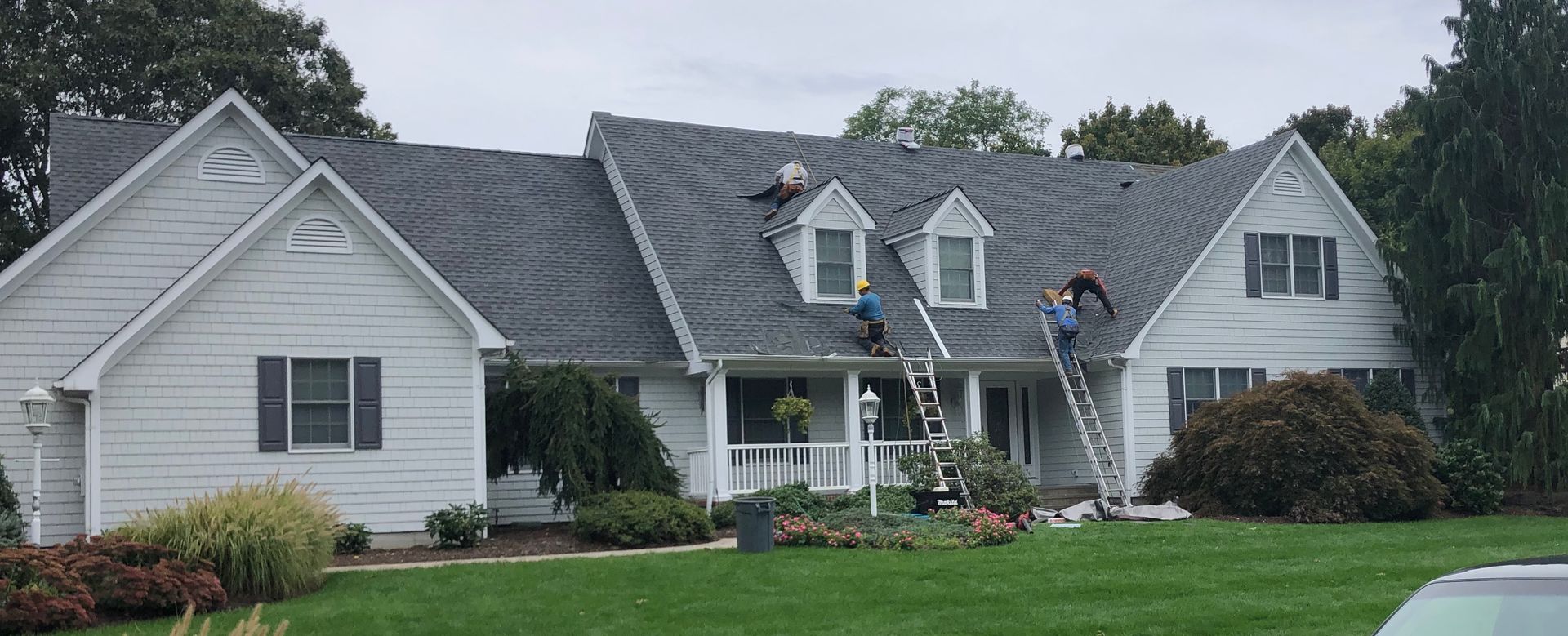 Workers on a roof installing shingles on a white house with two ladders propped against it