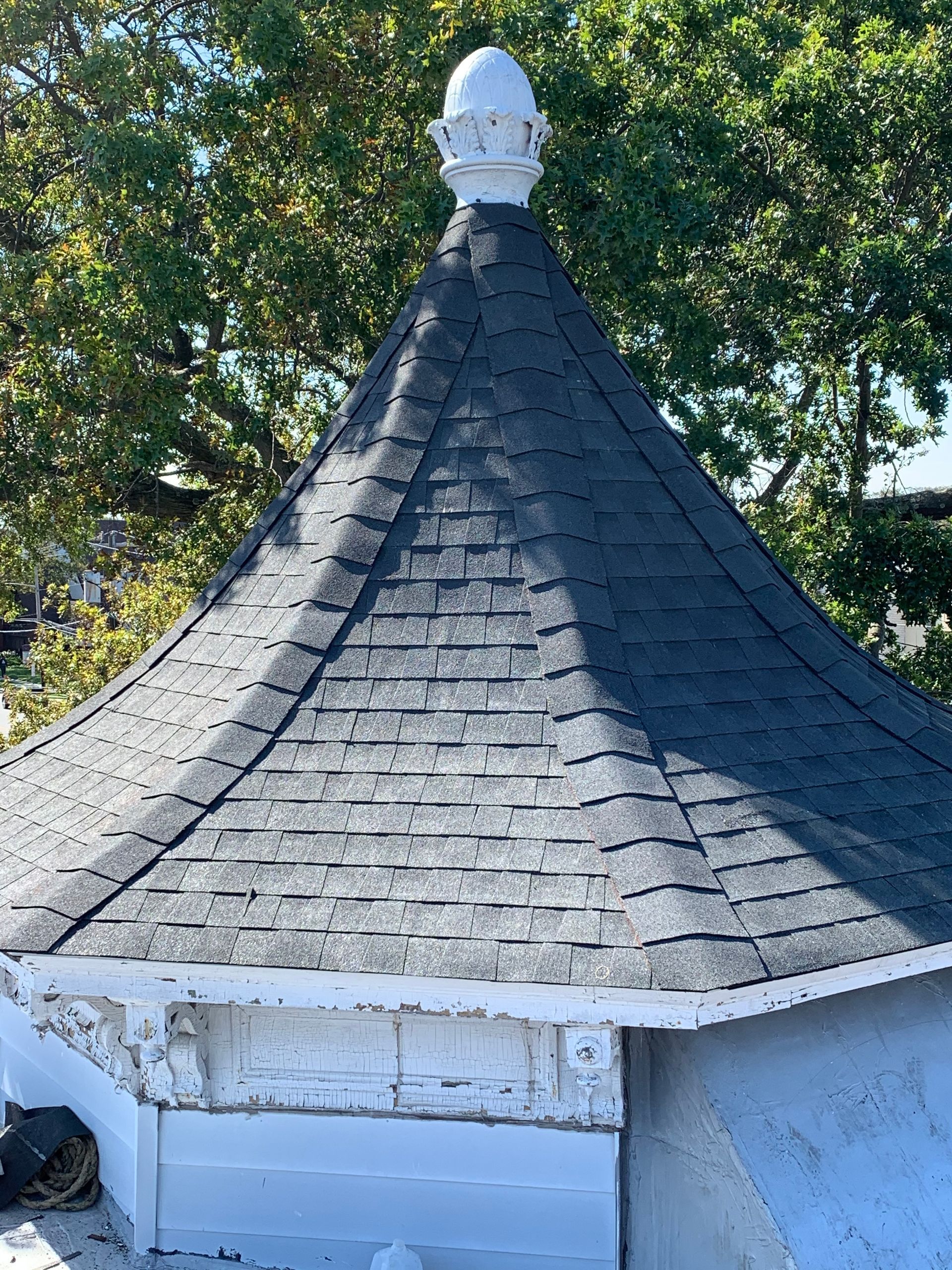 Dark shingled cone-shaped roof with a white decorative top against green trees