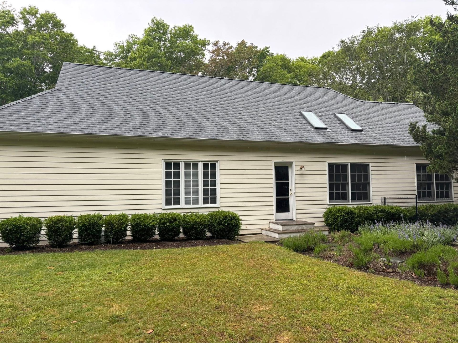 Single-story beige house with dark roof, white windows, and a front lawn surrounded by trees