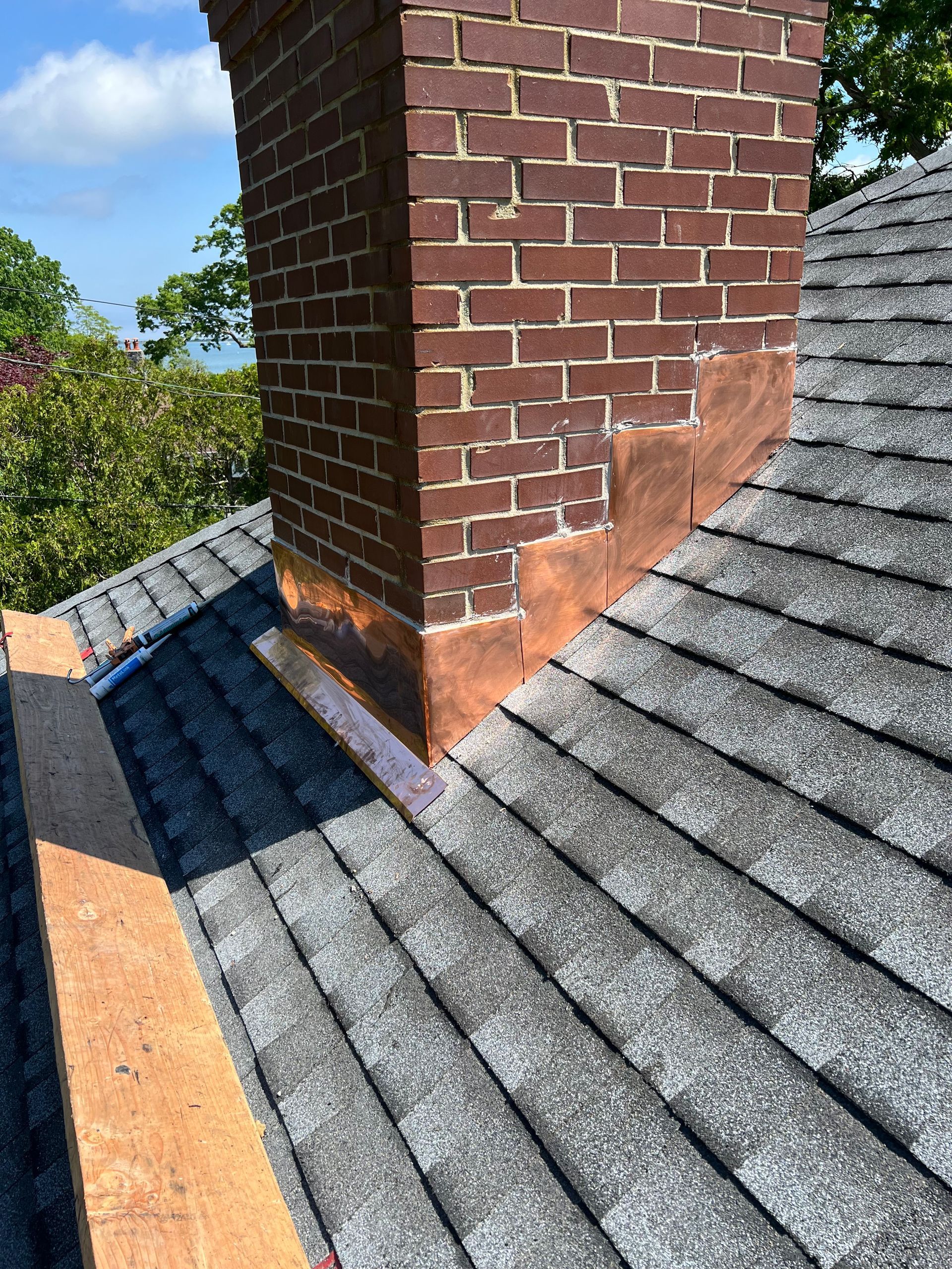Brick chimney on a shingled roof with copper flashing visible, cloudy sky and greenery