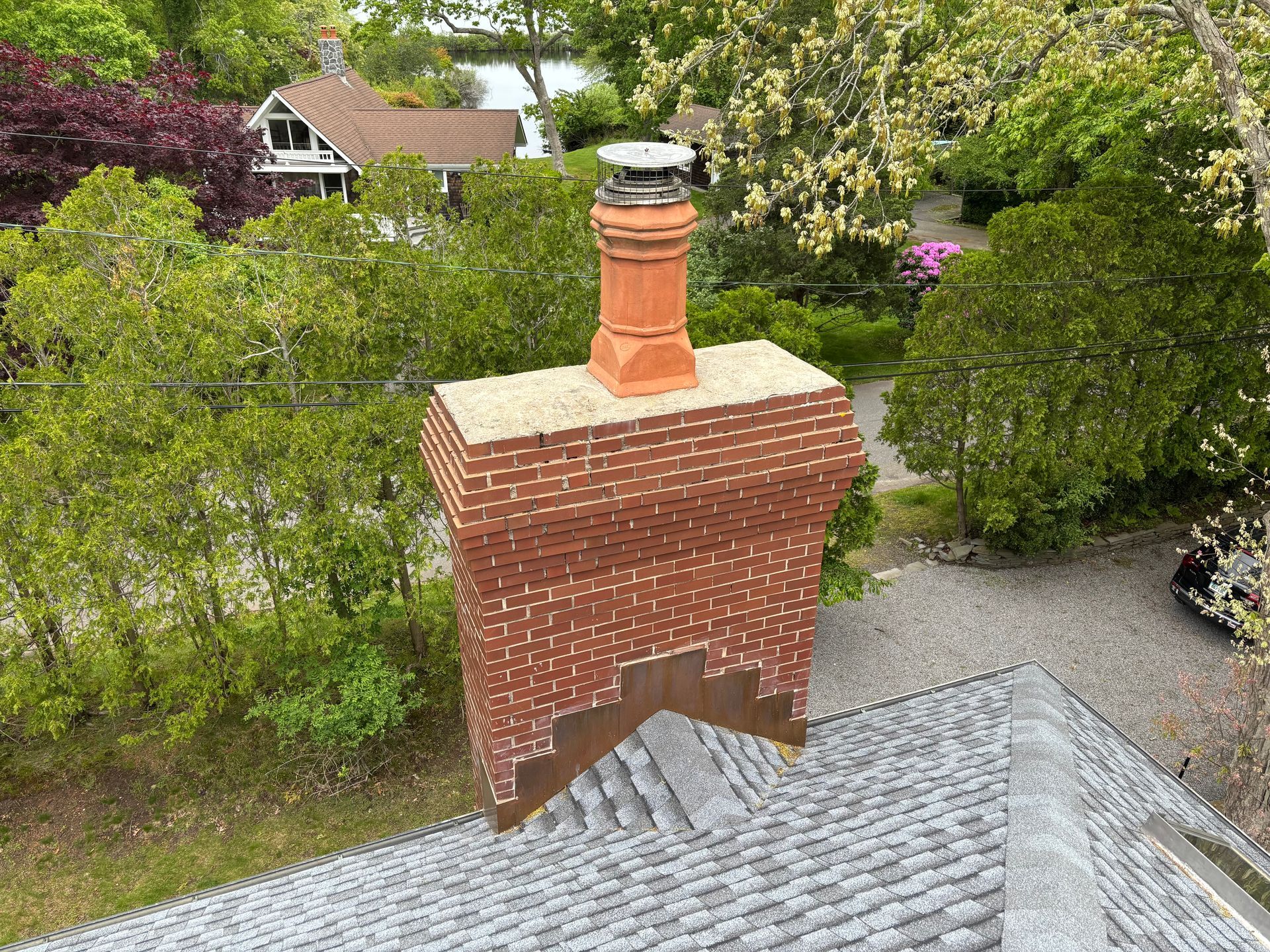 Brick chimney on a gray shingled roof topped with a terracotta flue and metal cap, surrounded by greenery