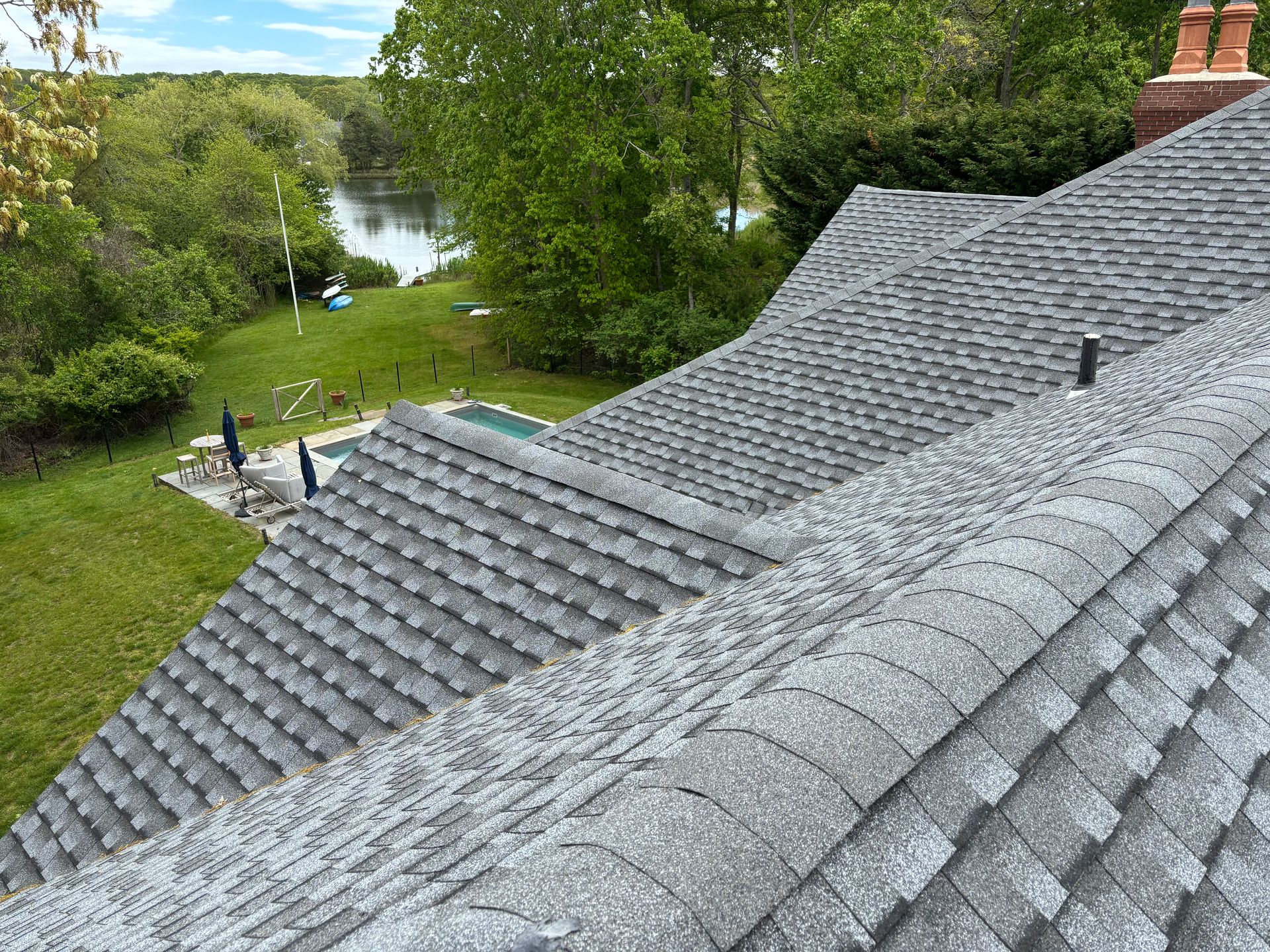 Gray shingled roof with a river and trees in the background, a swimming pool and patio below